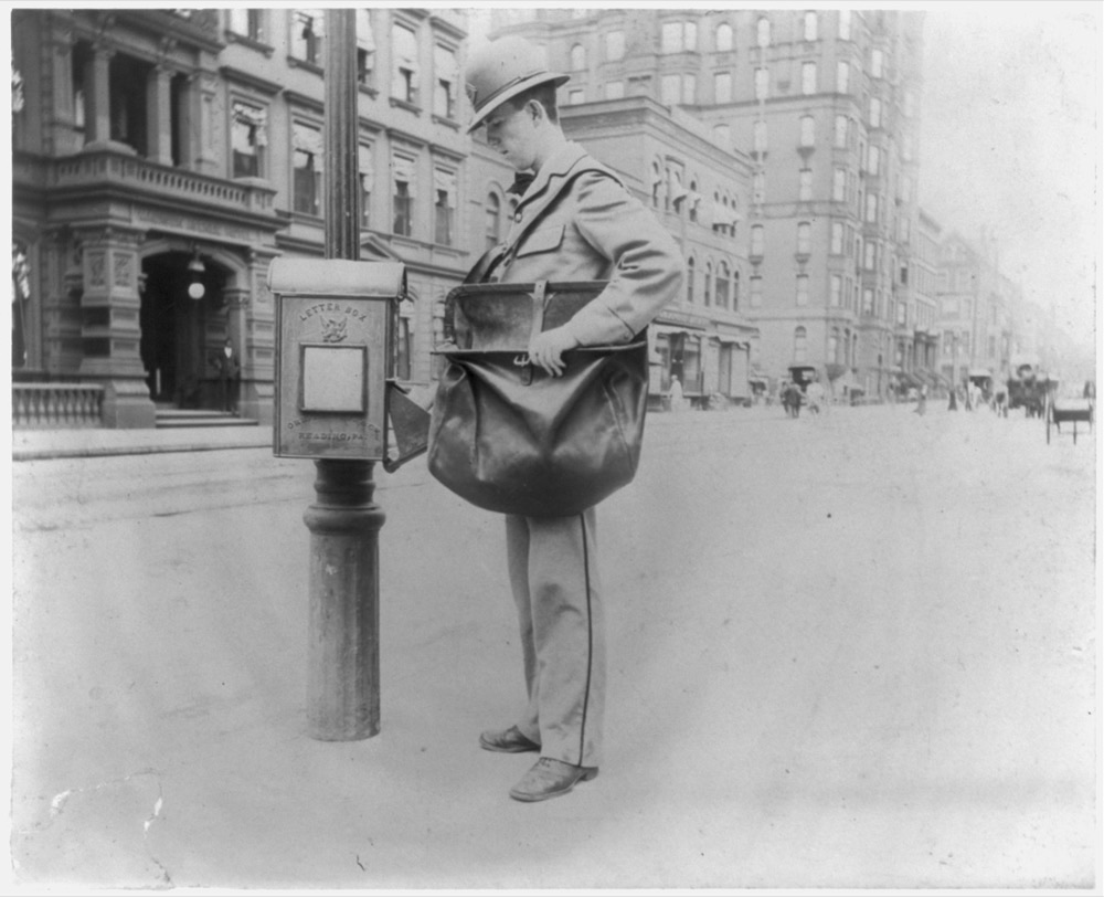 A late-19th-century New York City postman standing beside a street letter box, dressed in uniform.